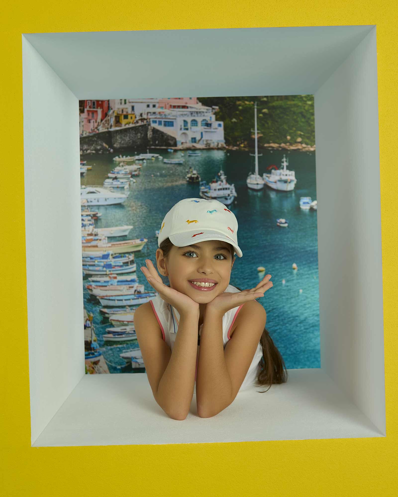 Bambina sorridente con cappellino bianco e sfondo marino con barche.