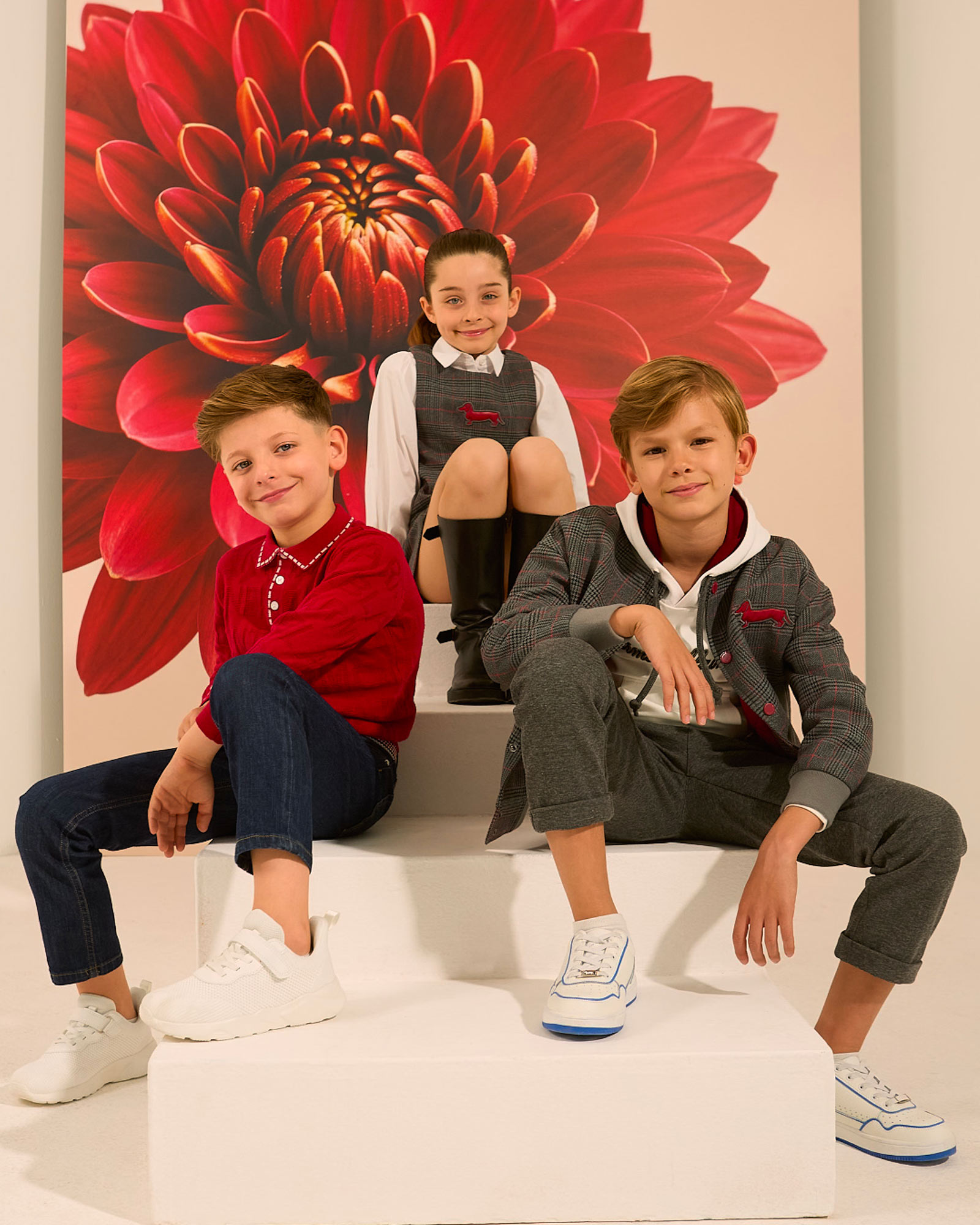 Three children in red and grey outfits with white sneakers in front of a large red flower.