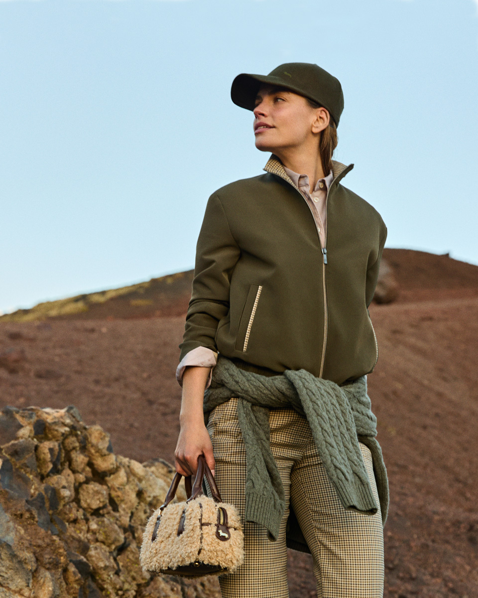 Female model wearing an olive green jacket, matching cap, patterned trousers, and a sweater tied around her waist, holding a small faux-shearling handbag against a rocky volcanic background.