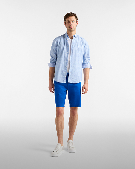 Full-length shot of a male model on a white background wearing bright electric blue chino shorts, paired with an unbuttoned light blue patterned shirt over a plain white t-shirt and white low-top sneakers.