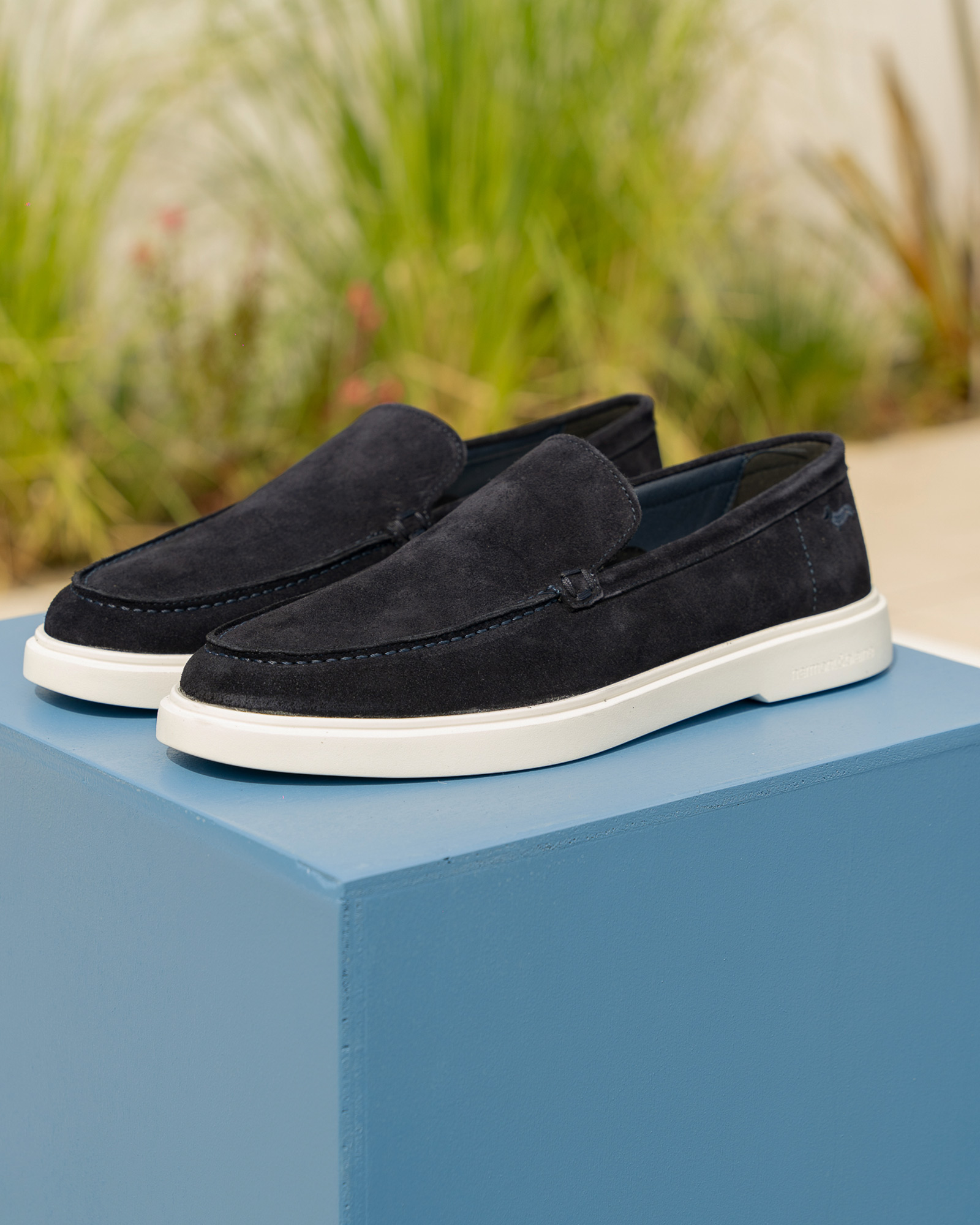 Men’s navy suede loafers with white sole, displayed on a light blue pedestal.
