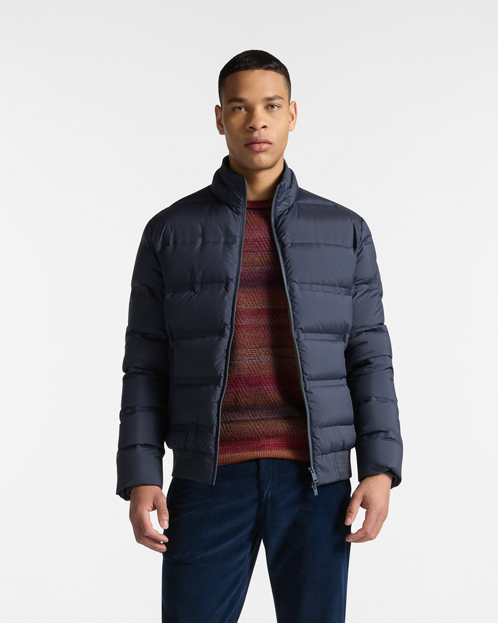 Hombre vistiendo una chaqueta acolchada azul oscuro con cremallera, un suéter de rayas multicolor y pantalones de pana azul. Fondo blanco neutro.